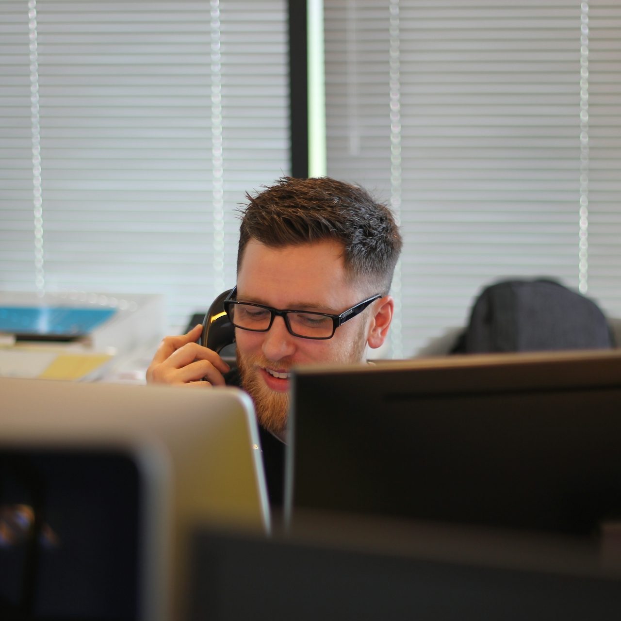 Mann mit Brille spricht am Telefon, sitzt am Schreibtisch vor Computerbildschirmen.